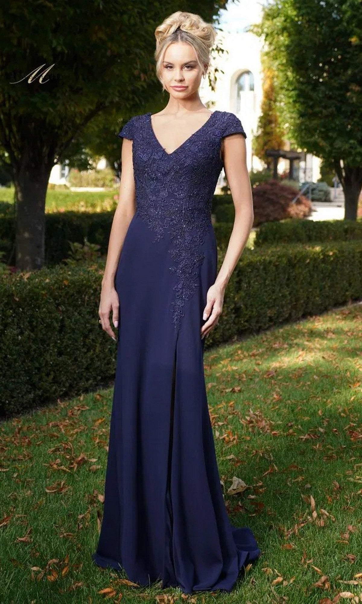 Outdoors on grass, a woman wears the Rina Di Montella RD2763 long formal dress in navy, featuring an embroidered bodice and short sleeves. Trees and shrubs are visible in the background.